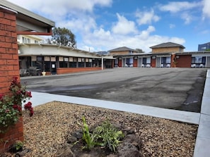 A hotel with a parking lot, a brick building on the left, and a row of rooms with windows. - The Abbotswood Motor Inn (Geelong)