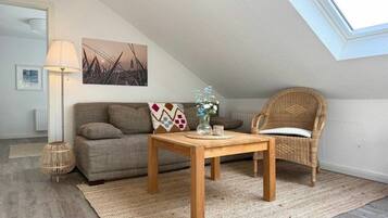 A cozy living room with a sofa, a wooden coffee table, a wicker chair, a lamp, and a painting on the wall.