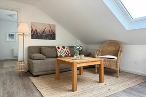 Ein gemütliches Wohnzimmer mit einer Couch, einem Holztisch, einem Korbstuhl, einer Lampe und einem Bild an der Wand.