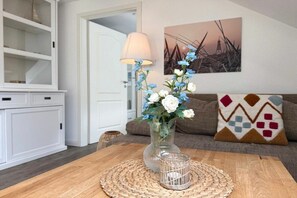 Ein Essbereich mit einem Holztisch, einer gewebten Tischmatte, einer klaren Glasvase mit Blumen, einer Lampe und einem Gemälde an der Wand.