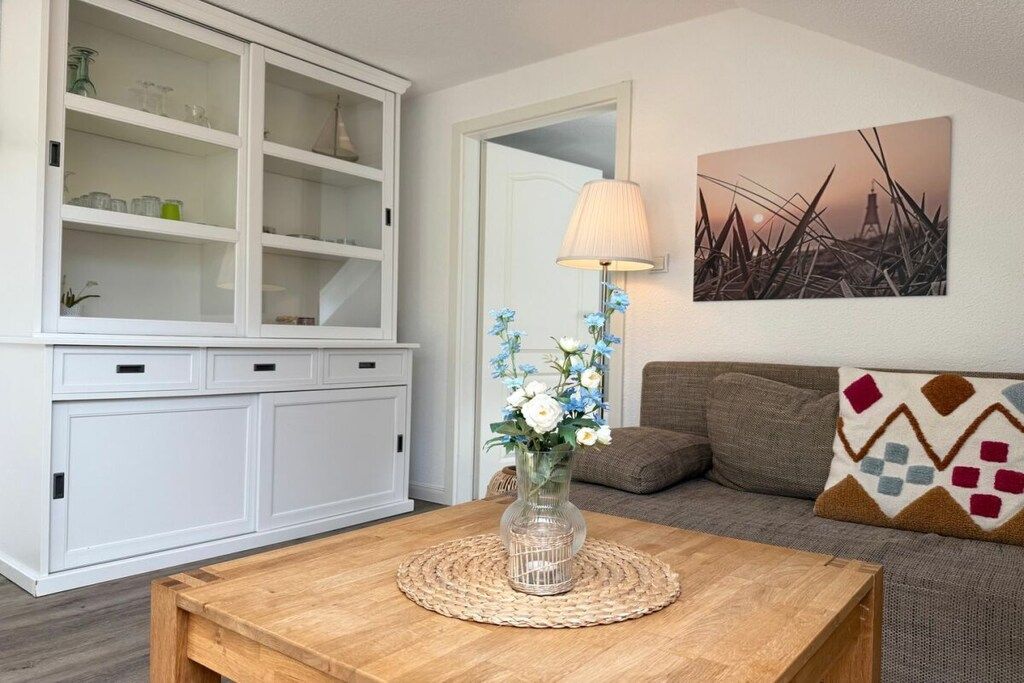 Ein Wohnzimmer mit einem Holztisch, einer weißen Vitrine mit Glastüren, einem Sofa mit gemusterten Kissen und einer Wandleuchte.