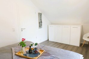 A bedroom with a bed, a bedside table with a tray of drinks and a vase of flowers, a white wardrobe, and a small white chair. - House No. 19, Fischerhus (Wangerland)