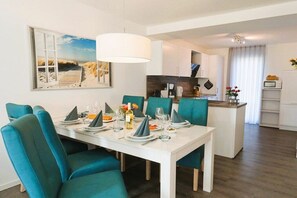 Uma sala de jantar com uma mesa branca posta para seis pessoas, cadeiras em tons de verde-água e um quadro com uma paisagem praiana.