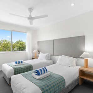 Habitación de hotel con dos camas, ventilador de techo, un ventanal y vista a árboles.