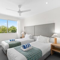 Habitación de hotel con dos camas, ventilador de techo, un ventanal y vista a árboles.