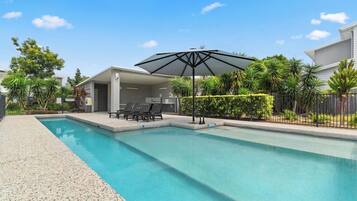 A swimming pool with a lounge area, a covered seating area, and a fence with greenery.