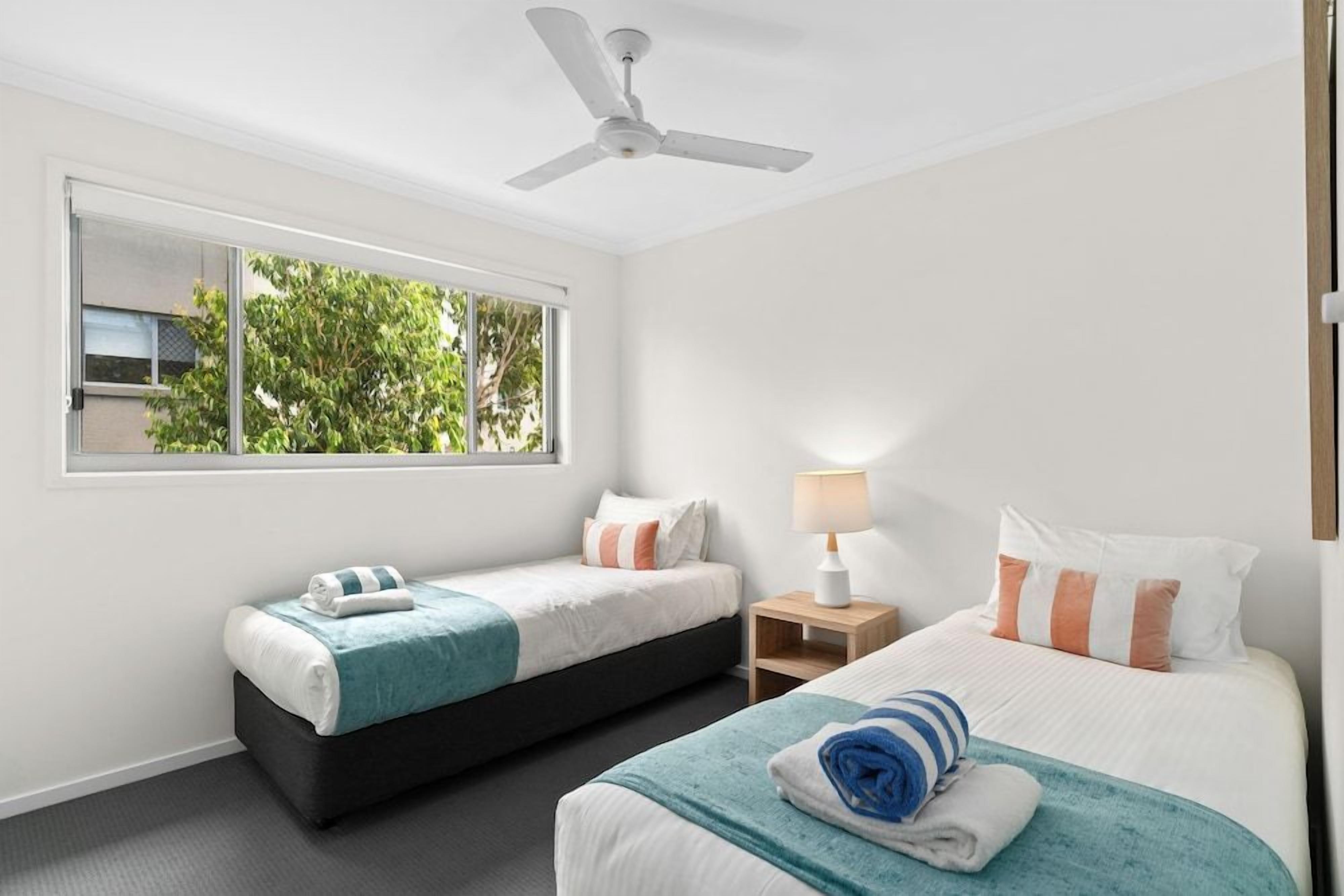 A hotel room with two beds, a ceiling fan, a window with a view of greenery, and a bedside lamp.