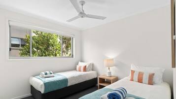 A hotel room with two beds, a ceiling fan, a window with a view of greenery, and a bedside lamp.