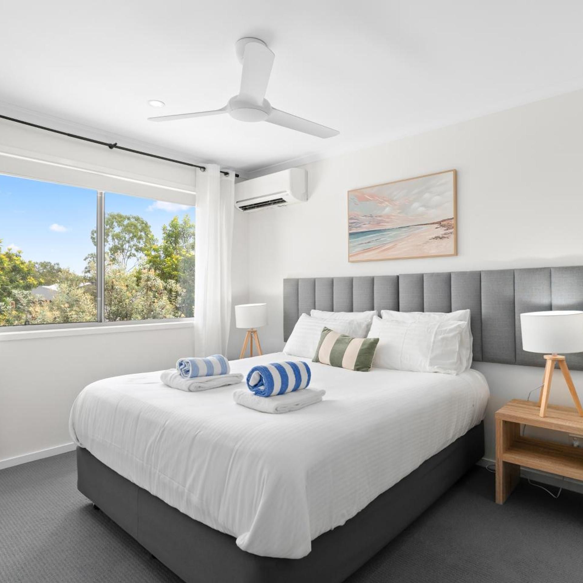 A modern bedroom with a large bed, a ceiling fan, a window with a view of trees, and a painting on the wall.