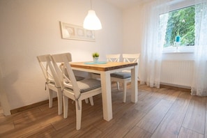 Ein Essbereich mit einem Holztisch, weißen Stühlen, einer Pendelleuchte und einem Fenster mit Vorhängen.