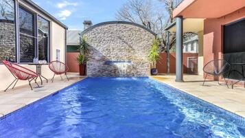 A swimming pool with a stone wall and a waterfall feature, surrounded by red chairs and potted plants.