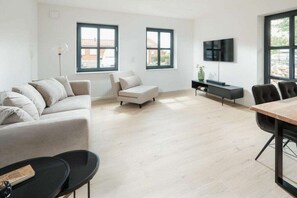 A modern living room with a sofa, a coffee table, a dining table with chairs, a TV, and two windows. - Coastal Suite South | Side Waterfront Location (Greetsiel, Germany)