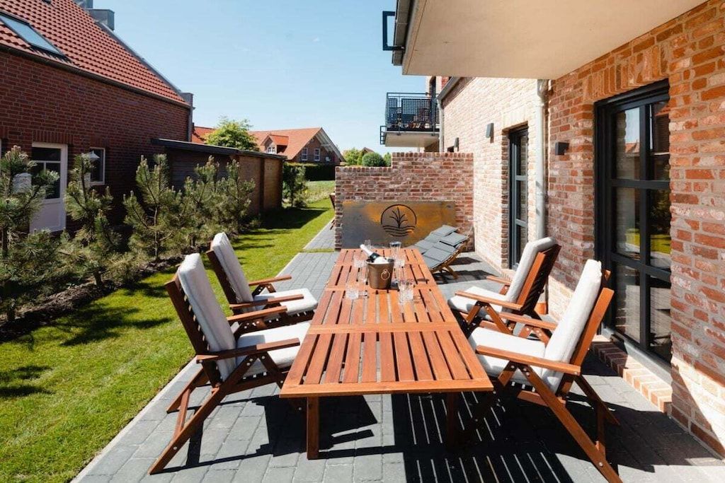 Un patio exterior con una mesa y sillas de madera, rodeado de muros de ladrillo y una zona ajardinada.