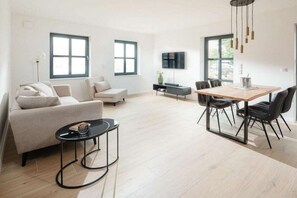 A modern living room with a sofa, a coffee table, and a dining area with chairs and a table. - Coastal Suite South | Side Waterfront Location (Greetsiel, Germany)