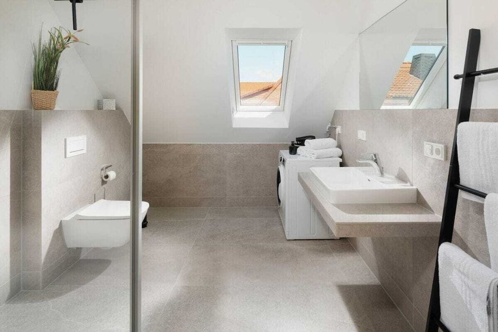 A modern bathroom with a toilet, bidet, and two sinks. There is a small window providing natural light and a view of the outside.