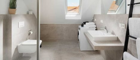 A modern bathroom with a toilet, bidet, and two sinks. There is a small window providing natural light and a view of the outside.