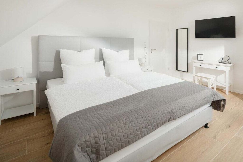 A modern bedroom with a large bed, white nightstands, a bedside table, a desk, and a wall-mounted TV.