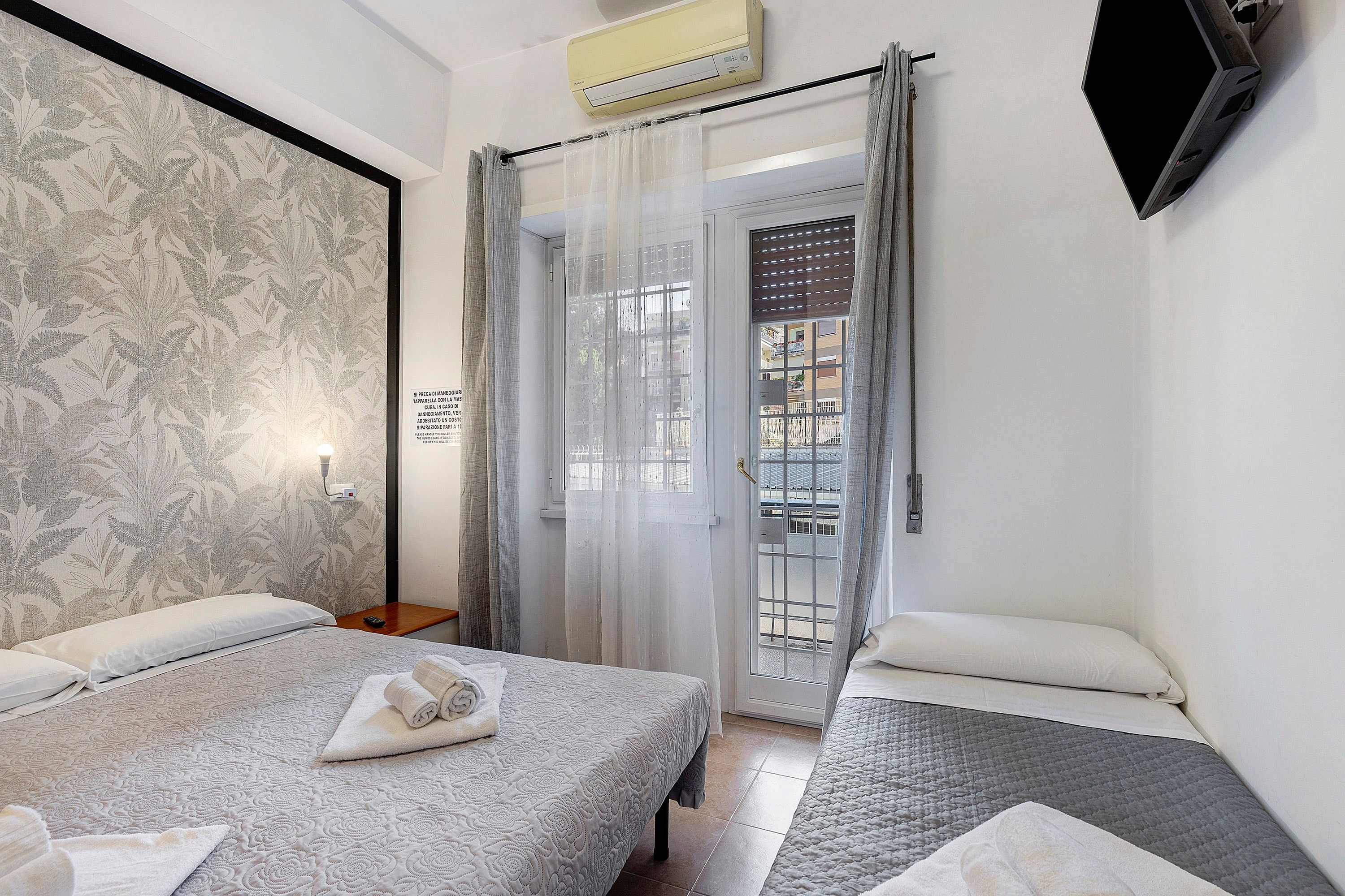 A hotel room with two beds, a large window with curtains, an air conditioning unit, and a wall-mounted TV.