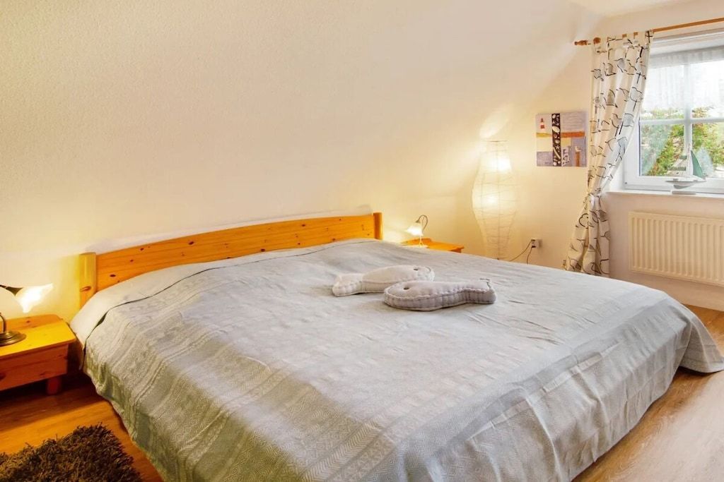 A bedroom with a wooden headboard, a bed with a grey bedspread, two slippers on the bed, a nightstand with a lamp, and a window with curtains.
