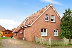 A two-story brick house with a sloped roof, a wooden fence, and a paved driveway. - Holiday home with 3 bedrooms (Neßmersiel)