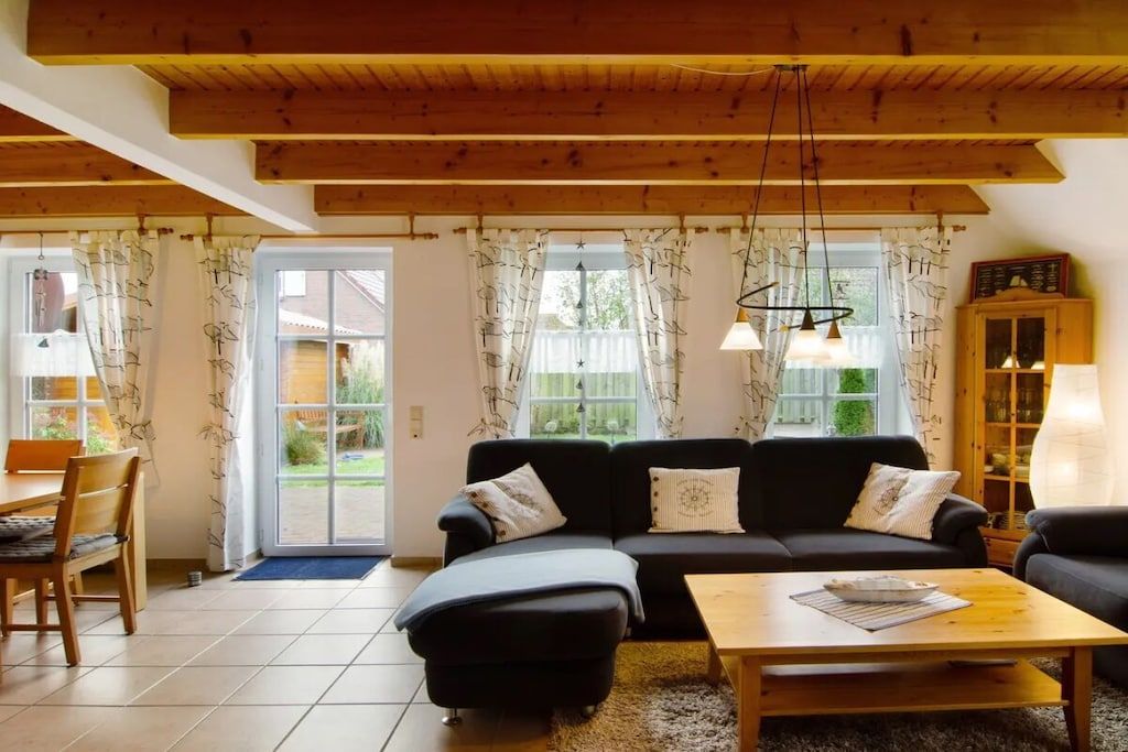 Una sala de estar con un sofá, una mesa de centro y un comedor con sillas. Hay una ventana con cortinas, una puerta que da al exterior y un techo de madera.