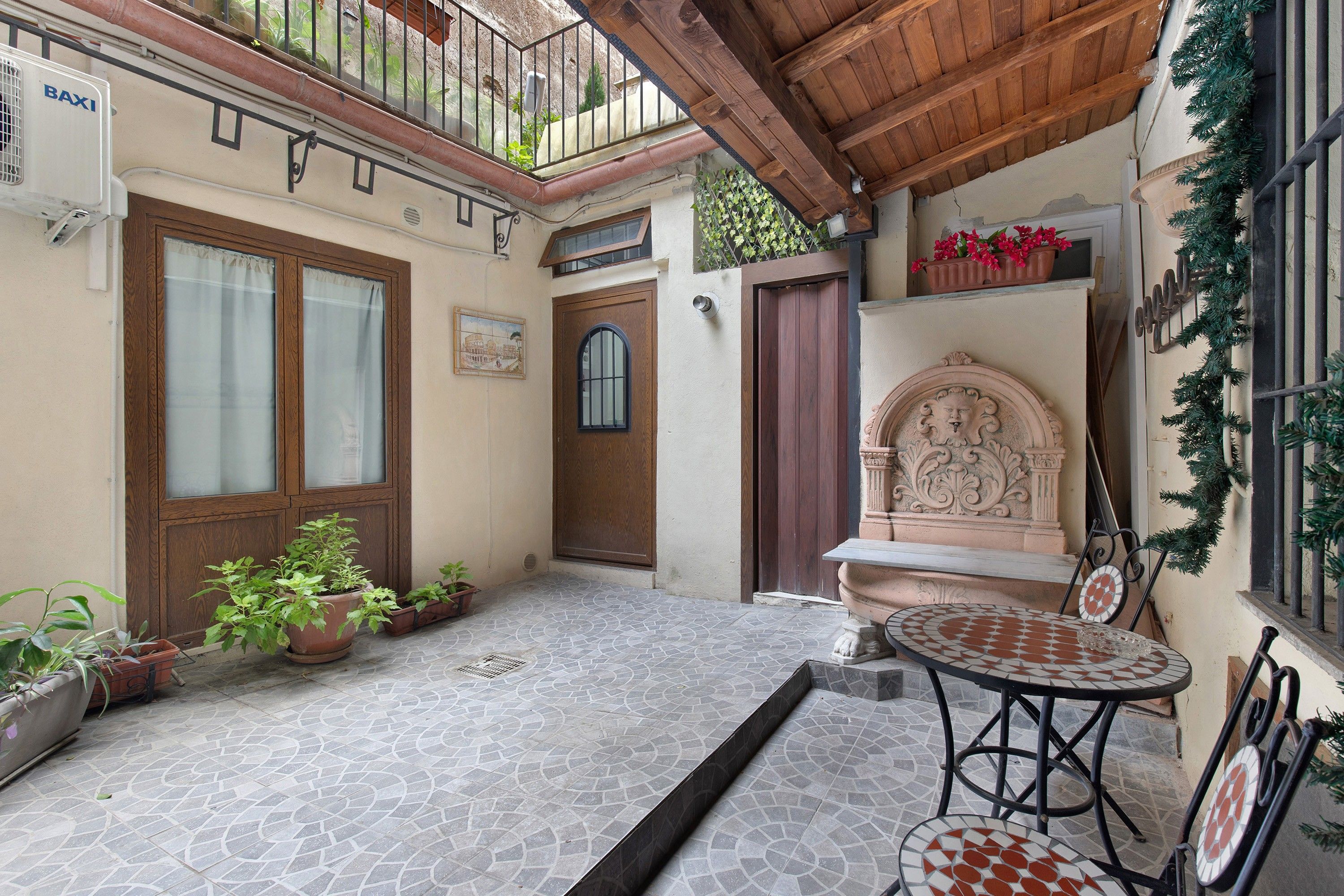 A courtyard with a tiled floor, a wooden door, a window with curtains, a potted plant, and a small table with chairs.