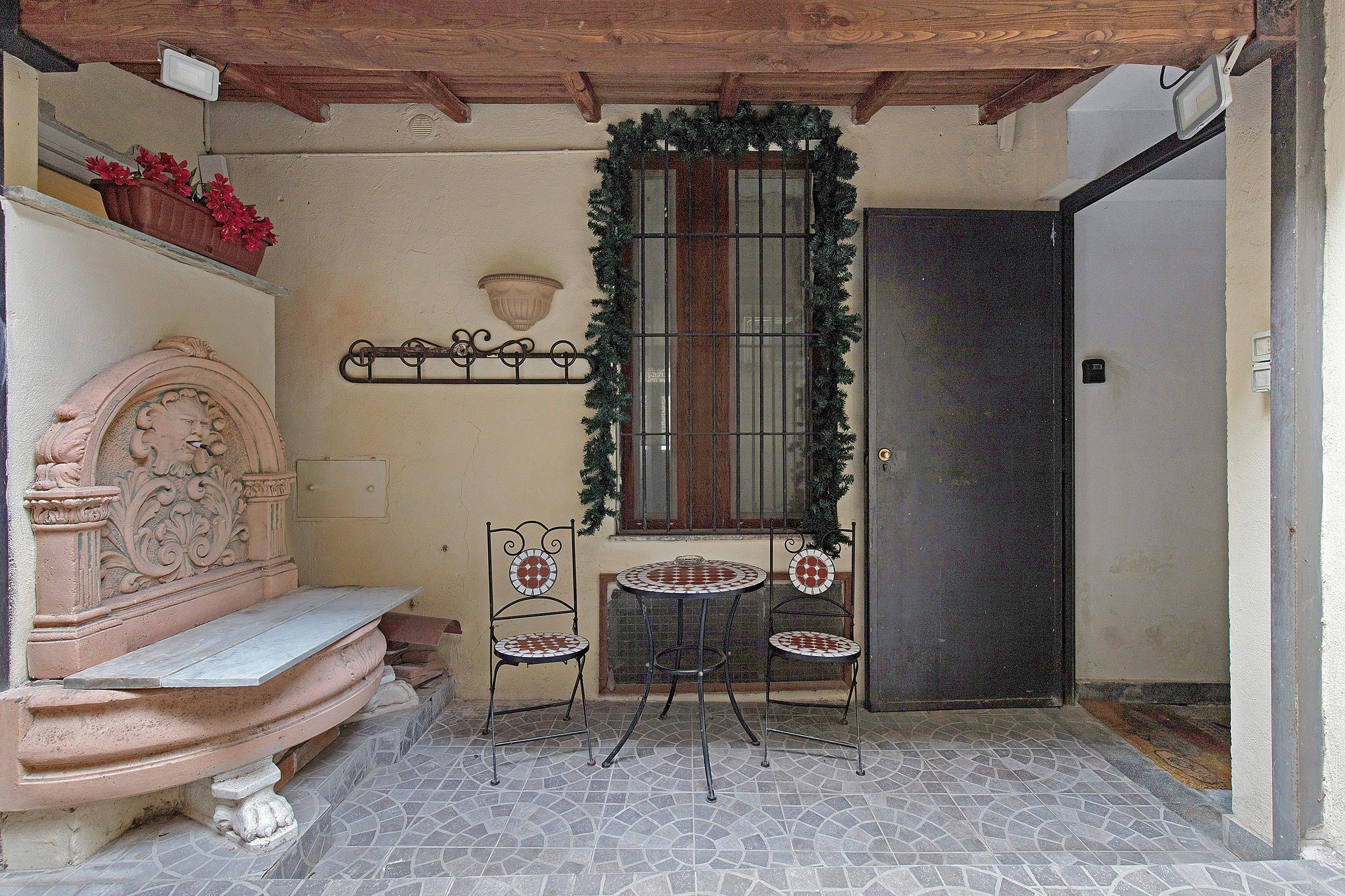 A small patio area with a bench, two chairs, a decorative wall, a window with a wreath, and a door.