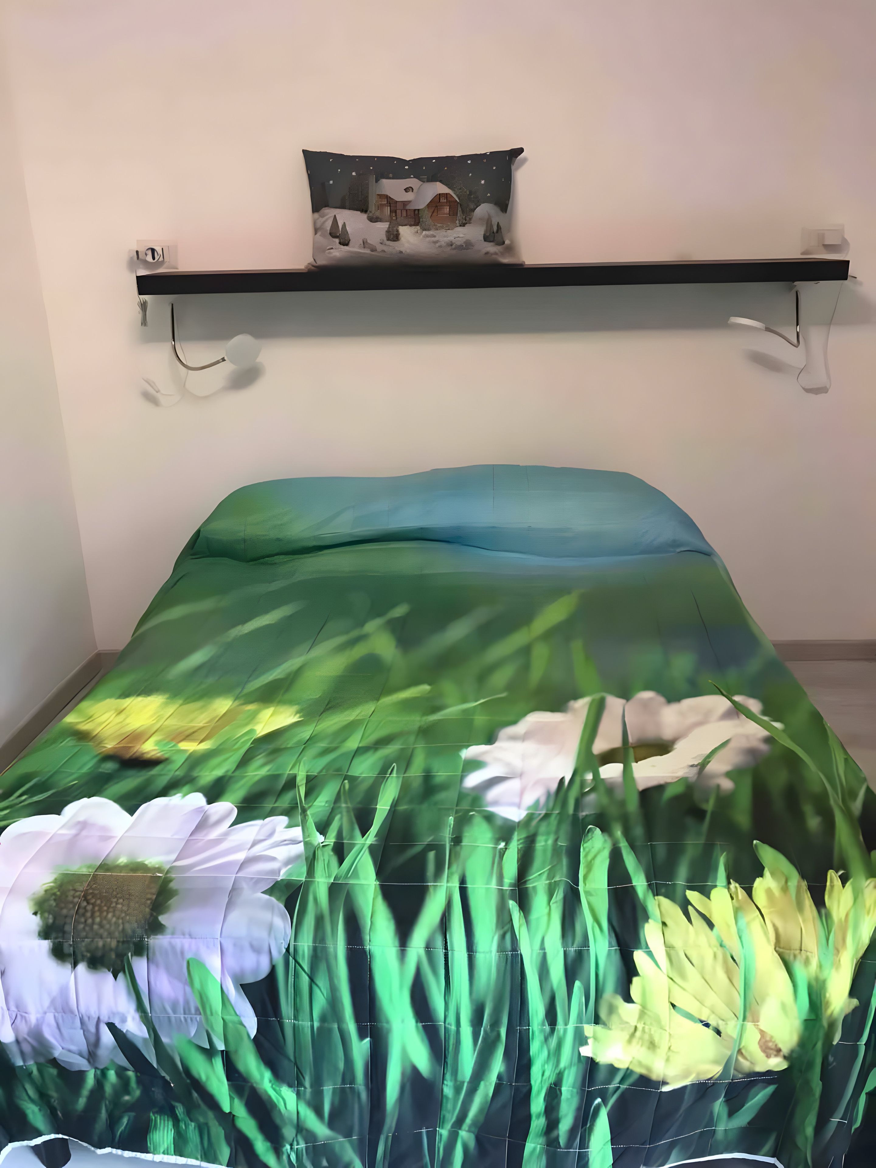 A bed with a floral bedspread, a bedside table with a lamp, and a wall-mounted shelf with a decorative item.