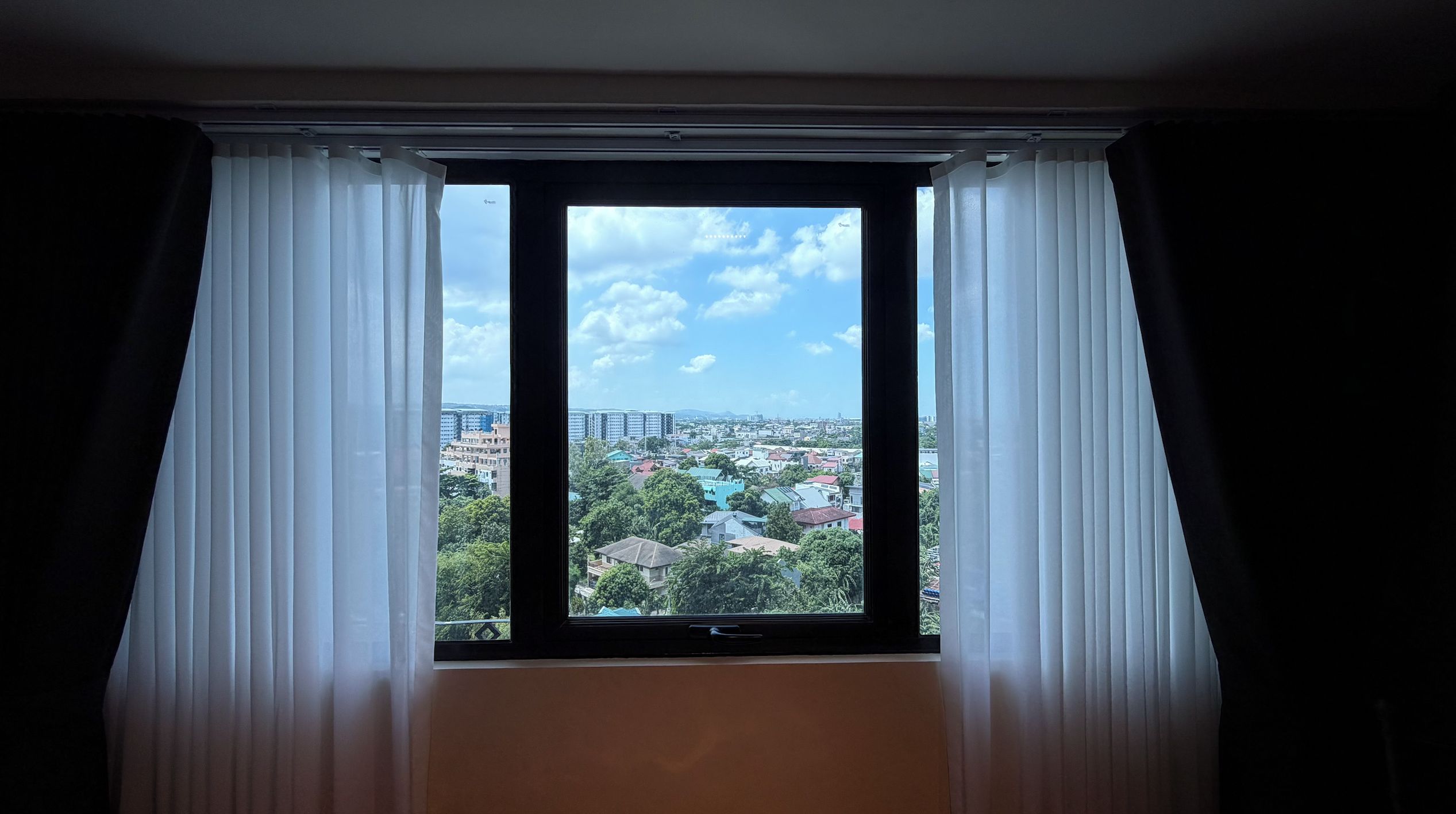 Una habitación con un ventanal grande y cortinas blancas, con vistas a un paisaje urbano.