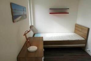 Ein kleines Zimmer mit einem Einzelbett, einem Holzkopfende, einem Nachttisch mit Lampe, einem Wandregal und einem Gemälde.