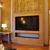 Una sala de estar moderna con un televisor de pantalla plana grande montado en una pared de madera con patrones geométricos, una mesita con una lámpara y suelo de madera.