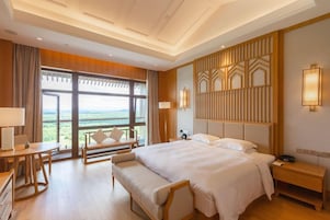 A hotel room with a large bed, a desk, a bench, and a view of the outdoors.
