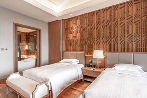 Habitación de hotel con dos camas, cabecera de madera, mesita de noche con lámpara y espejo.