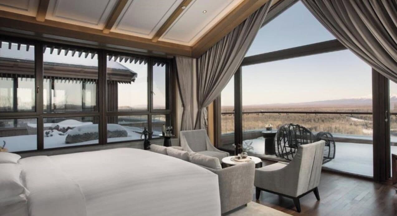 Een slaapkamer met een groot bed, een bank, een klein tafeltje en stoelen. Er zijn grote ramen met gordijnen, en een uitzicht op een besneeerd landschap.