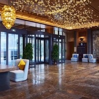 A lobby with a modern design, featuring a large chandelier, a reception desk, seating area, and a glass door entrance.