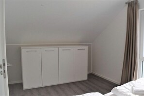 Une pièce avec une armoire blanche, un lit avec une literie blanche et une fenêtre avec des rideaux.