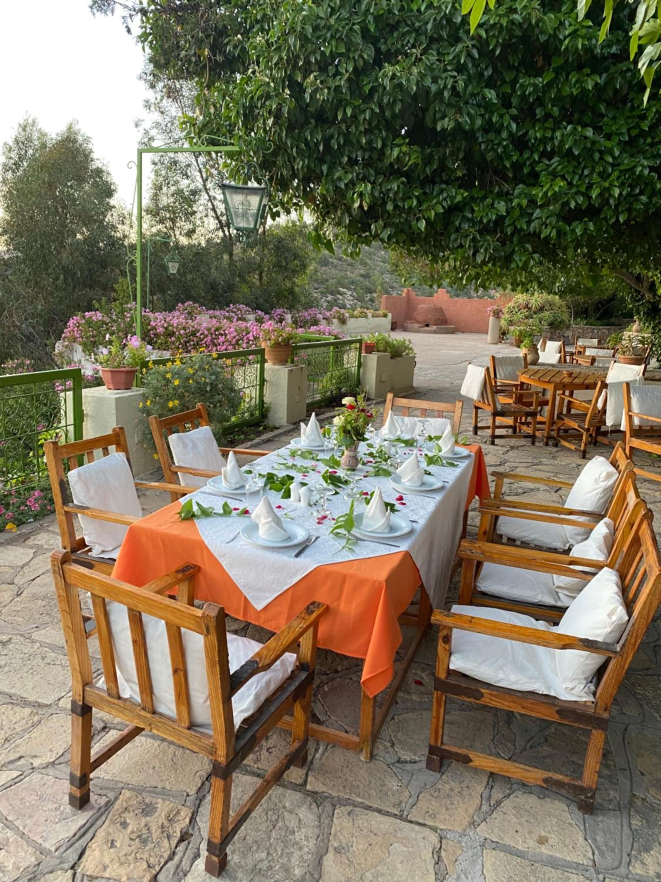 An outdoor dining area with stone flooring, wooden tables, and chairs, surrounded by greenery and potted plants.