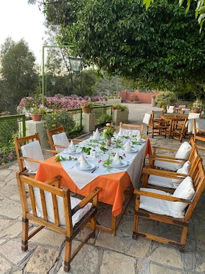 An outdoor dining area with stone flooring, wooden tables, and chairs, surrounded by greenery and potted plants.