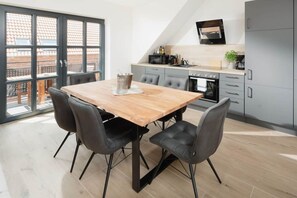 A modern kitchen with a dining area, featuring a wooden table and black chairs, a built-in oven, and a TV mounted above the counter. - Coastal Suite West | Side Waterfront Location (Greetsiel)