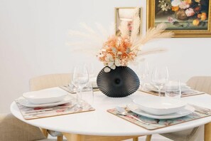 Ein runder Esstisch mit weißen Tellern, klaren Gläsern und einer schwarzen Vase mit getrockneten Blumen.