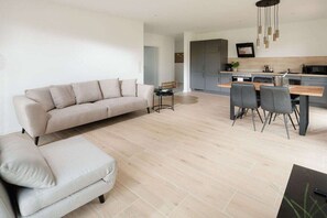 Een moderne woonkamer met een bank en voetenbankje, een eethoek met stoelen en een tafel, en een keuken met kasten en apparatuur.
