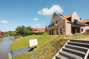 A modern building with a stone facade, a balcony, and a grassy area with a wooden deck leading to a body of water. - Coastal Suite North | Directly on the waterfront (Greetsiel)