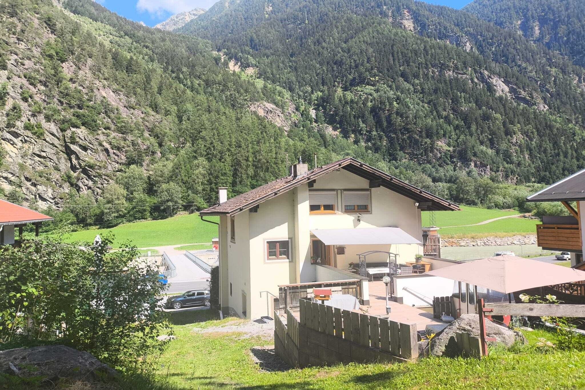 Une maison à deux étages avec un balcon, entourée d’une cour clôturée et dominant un paysage montagneux.