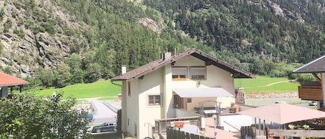 Une maison à deux étages avec un balcon, entourée d’une cour clôturée et dominant un paysage montagneux.