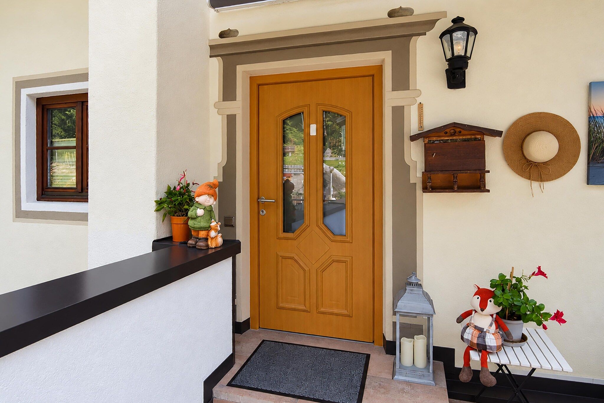 Une porte d’entrée en bois avec un panneau de verre, un rebord noir, une petite fenêtre, une lanterne fixée au mur, un panier en osier et une figurine décorative avec une plante.
