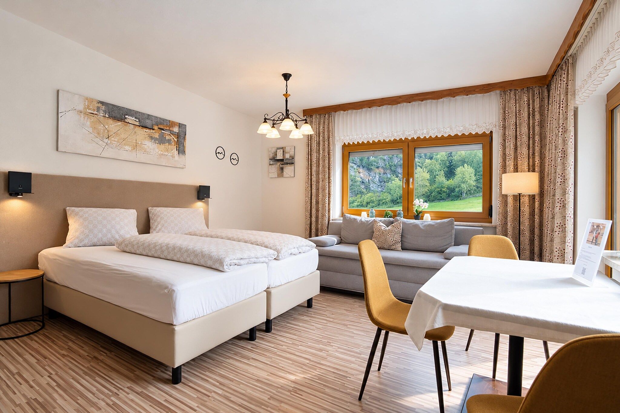 A hotel room with a double bed, a sofa, a dining table, and a view of greenery through the windows.