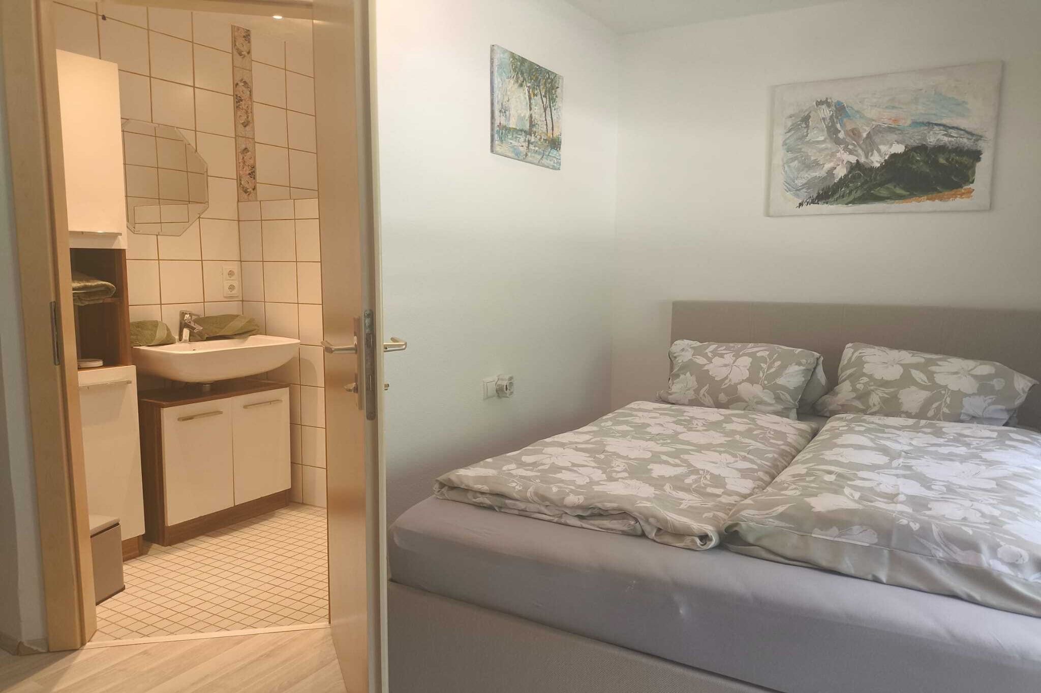 A bedroom with a bed, a nightstand, a wall-mounted painting, and a bathroom with a sink and mirror.
