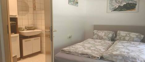 A bedroom with a bed, a nightstand, a wall-mounted painting, and a bathroom with a sink and mirror.