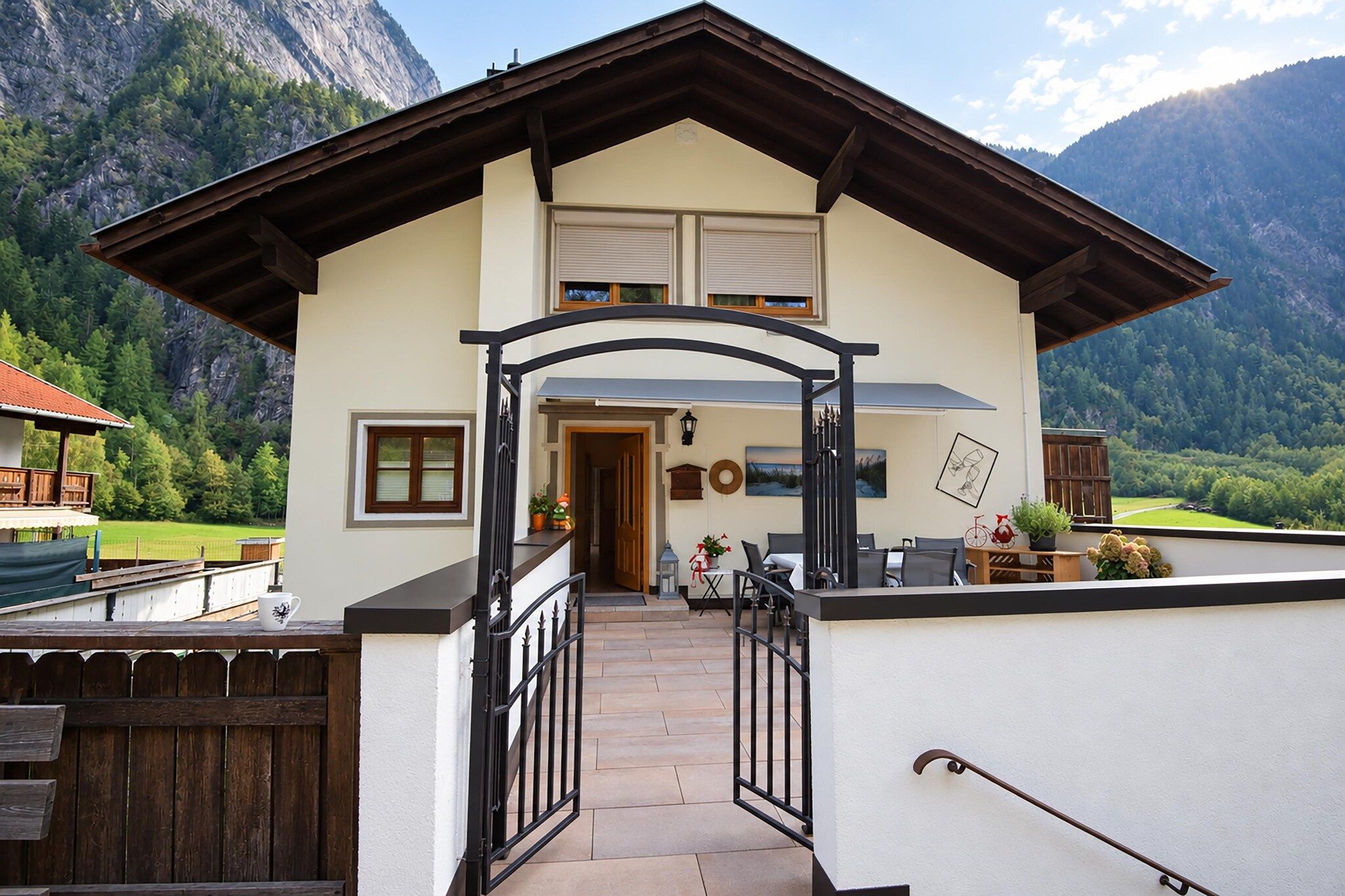A two-story chalet with a balcony, a wooden deck, and a mountain backdrop.