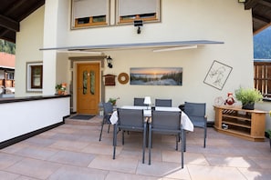 Een terras buiten met een tafel, stoelen, een houten deur en een aan de muur bevestigd schilderij.